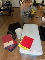 Photo showing large and small storage bins, trash bin, flatware storage container, and photo/video storage boxes placed on the floor along with a red spiral notebook.