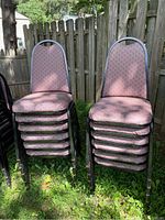 Twelve stacking chairs grouped in two stacks outdoors against a wooden fence. Chairs have black metal frames and pinkish patterned fabric upholstery. Rust visible on frames and fabric stains visible.