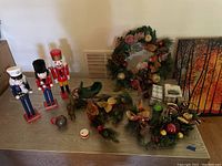 Photo showing three nutcracker figurines in red, blue, and black/white, multiple Christmas wreaths decorated with pine cones, glittered and colored balls, and leaves, along with a small green sleigh and a metal donkey figure on a wooden table.