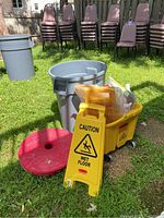 Photo of a yellow wet floor caution sign, yellow rolling mop bucket, gray trash can, and stack of chairs in background on grass.
