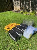 Image showing multiple rectangular plastic storage bins with black lids, several clear plastic containers with yellow lids, a yellow hanging storage organizer, and a metal wire basket on grass.