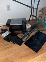 Image showing Canon MP390 printer, three black Samsung tablets, RCA notebook computer, two black TV mount bases with metal stands, and power cords on wooden floor near wall outlet.