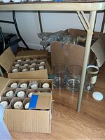 Box of glass candle holders filled with white round wick candles, placed on floor beneath table.