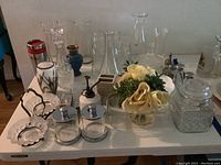 Wide shot of assorted glassware on white table including vases, decanter, glass jars, artificial flower arrangement, and other glass items.