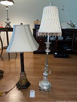 Two lamps on wood floor, one with clear glass base and white beaded shade, other with twisted metal stem and beige shade