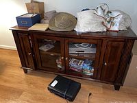 Front view of the wood media cabinet showing glass doors and contents inside shelving. Items on top are textiles and a hat, not included.