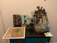 Photo showing the full set of items on a teal dresser, including the Lenox Falkenstein castle replica, glass decanter, wooden trinket box, metal piano player figurine, decorative plaques, and letter B plaques.