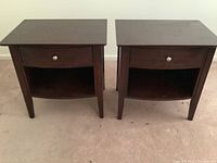 Front view of the pair of brown stained wooden side tables showing one drawer with chrome knob and lower shelf.