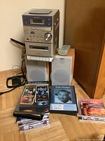 Sony disc deck receiver stacked atop two Sony bookshelf speakers with a remote on one speaker and multiple CDs, DVDs, and cassettes arranged in front on the floor.