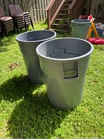 Two gray plastic trash cans standing upright on grass, showing overall shape and size