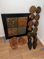 Full set of art pieces arranged on floor against wall: large framed metal embossed art, vertical multi-piece circular metal floral art, two wooden masks, and two leather mask sculptures.