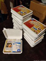 Photo showing 10 stacked white plastic auto first aid kits each labelled and compact, with one kit opened displaying packaged poncho and combine pad inside.