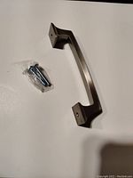 Metallic nickel finished cabinet/drawer pull lying flat with two mounting holes visible and accompanying screws in a plastic bag.