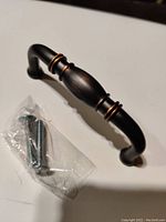 Single bronze patina drawer pull handle with two mounting screws beside it on a white surface, showing overall design and color.