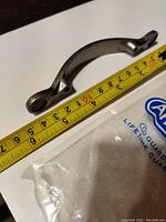 Close-up side view of aged nickel cabinet pull next to measuring tape showing length around 4 inches.