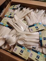 Box filled with multiple clear-packaged sets of white plastic cutlery including spoons, forks, and knives.