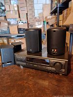 Front view of the Technics amplifier with two Infinity Micro II speakers and one speaker stand mount on a brown surface, in a room with many stacked boxes.