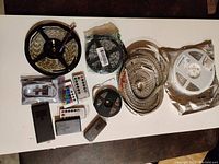 Photo showing multiple LED strip light reels and coiled bundles along with several infrared remote controls and control units on a white surface