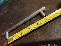 Close-up photo of a single long bar-style cabinet handle in brushed nickel finish showing a measuring tape indicating a 5-inch distance between screw holes.