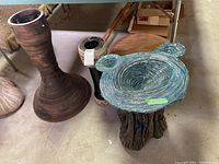 Photo showing the bird bath with blue-green textured basin and sculpted base, large brown planter base, and smaller cream-colored planter base with price tag, all placed on concrete floor.