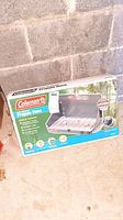 Original boxed Coleman two burner propane stove in unopened packaging with product labeling showing features and brand.