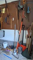 Wide view of pegboard wall showing multiple garden and hand tools hanging including shovels, rakes, stakes, and screwdrivers.