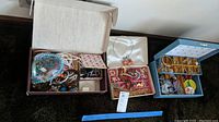 Three boxes open showing assorted costume jewelry including beaded necklaces, bracelets, bangles, sunglasses, earrings, and pins.
