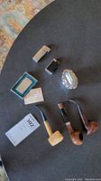 Top-down view of the three pipes and three lighters arranged on a black surface, including boxed Zippo lighter and paperwork.