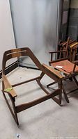 Wooden Mid-Century Modern rocking chair frame with armrests, showing general wear and scratches, situated on concrete floor with directors chairs in background.