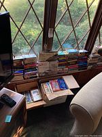 Wide view of multiple stacks and boxes of paperback books near a window, showing titles and arrangement.