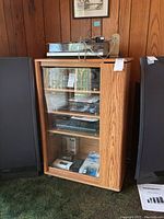 Front view of wooden stereo cabinet against wood panel wall with large speakers on either side. Glass door shows contents inside including audio equipment and accessories.