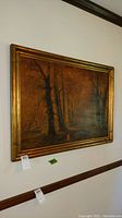 Framed painting showing an autumn forest with tall trees and fallen leaves, gold-toned decorative frame.