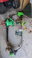 Green Machine 31cc weed whacker and Expert Gardener 2-speed blower vac on garage floor with rake partially visible in background.