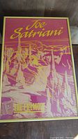 Framed poster with Joe Satriani text and Silver Surfer themed artwork in pink and yellow tones, viewed front on.