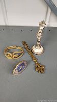 Four trinkets displayed on a gray surface including a gold rope bracelet, a floral enamel ring, an ornate letter opener, and a silver hand paper clip stand.