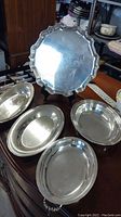 Five silver serving platters displayed on a wooden table, showing varying shapes and designs including one large round scalloped platter and four oval trays.