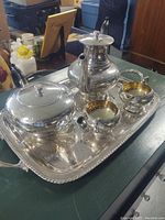 Photo showing the full set of 5-piece silver tea set on a rectangular tray with reflective surfaces and gold-toned interiors on bowls.