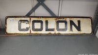 Full view of the rectangular metal street sign with 'COLON' in black letters on white background, showing age wear and rust.