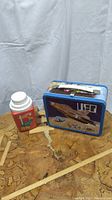 Photo showing the blue metal lunch box featuring a UFO spaceship graphic and the red Hot Wheels thermos with screw-on cap.