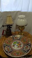 Lot arranged on wooden surface showing all three main items: oil lamp with glass shade, Asian plate, and blowtorch lamp with small lampshade.