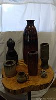 Photo showing seven pottery pieces on a wooden slab including the large dark brown vase and other smaller vases and jug.