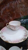 Photo of a delicate white tea cup with pink floral design and gold-trimmed handle resting on a matching plate with scalloped pink and gold rim.
