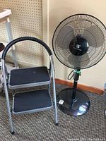 Full view showing the folding step stool next to the oscillating floor fan on a carpeted floor.