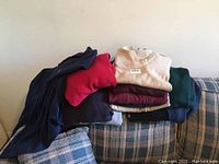 Stack of men's sweaters in various colors including cream, burgundy, navy, red, green, black on a sofa.