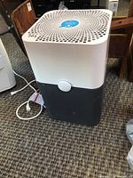 Front and angled view of the Blueair Pure 211+ air purifier showing white top with perforated holes and dark blue bottom with central button.