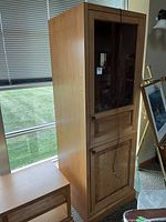 Side view of tall wooden display cabinet with glass front door, metal handle, and pull-out drawer below