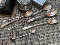 Various electroplated serving cutlery including spoons, forks, and ladles with ornate and dated markings on handles laid on carpet.