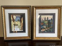 Both framed artworks showing street scenes in gold frames, side by side on a table.