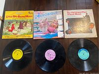 Three vintage children's vinyl LP records laid out with their sleeves on a wooden floor, showing front cover art for Disney's Little Red Riding Hood and Other Best Loved Fairy Tales, Miss Piggy Aerobique, and The Christmas That Almost Wasnt.