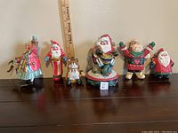 Full view of seven detailed wooden Christmas ornaments arranged in a row, including various Santa Claus figures and holiday characters.